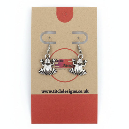 A pair of silver-toned frog dangle earrings displayed on a kraft paper card with red accents.