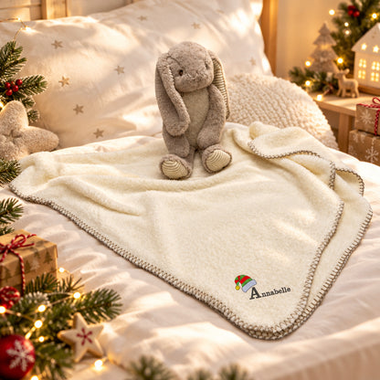 A personalized cream baby blanket with a knit border and an embroidered 'Annabelle' name with an elf hat, styled on a bed with a plush bunny toy, holiday decorations, and warm string lights.