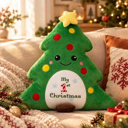 A plush Christmas tree-shaped pillow with embroidered facial features and the words 'My 1st Christmas' on the front, styled on a cozy sofa with holiday decorations and warm blurred lights in the background.