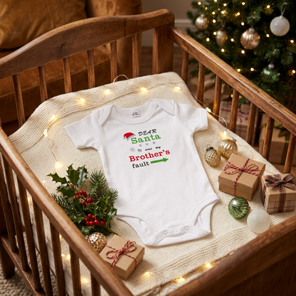 A white baby bodysuit with a Christmas-themed graphic reading 'DEAR Santa it was my Brother's fault' lies on a cream knit blanket inside a wooden crib. The scene is styled with holiday greenery, wrapped gift boxes, string lights, and a blurred Christmas tree in the background.