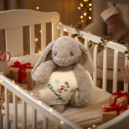 A plush gray bunny stuffed animal sits in a white crib decorated for Christmas, holding a cream pillow embroidered with 'My First Christmas.' The scene includes wrapped gift boxes, candy canes, string lights, and holiday garlands in a cozy nursery setting.