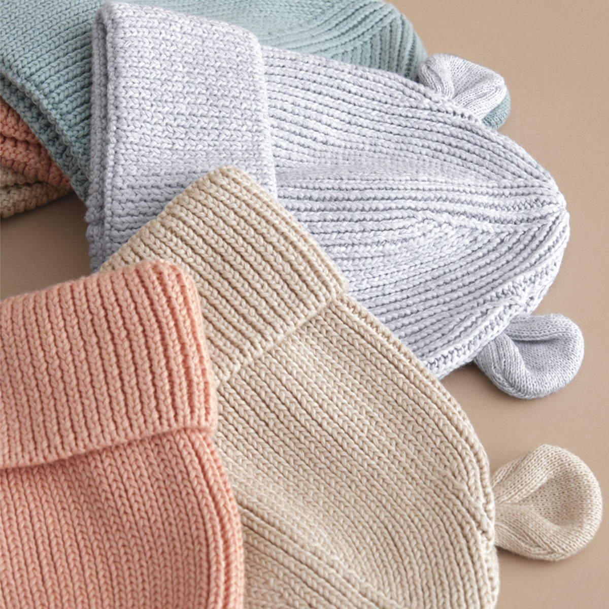 Four ribbed knit baby hats in soft pastel colors, including dusty pink, cream, light blue, and mint green, arranged on a beige background.