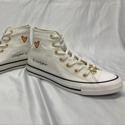 A pair of white high-top canvas sneakers with gold eyelets, gold heart details on the ankle, and gold signature charms on the laces, displayed on a wrinkled white fabric background.
