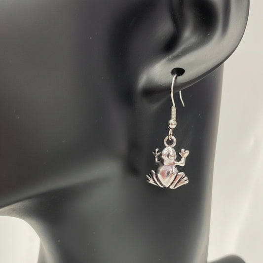 A silver-toned dangle earring with a small frog charm displayed on a black mannequin head against a white draped fabric background.