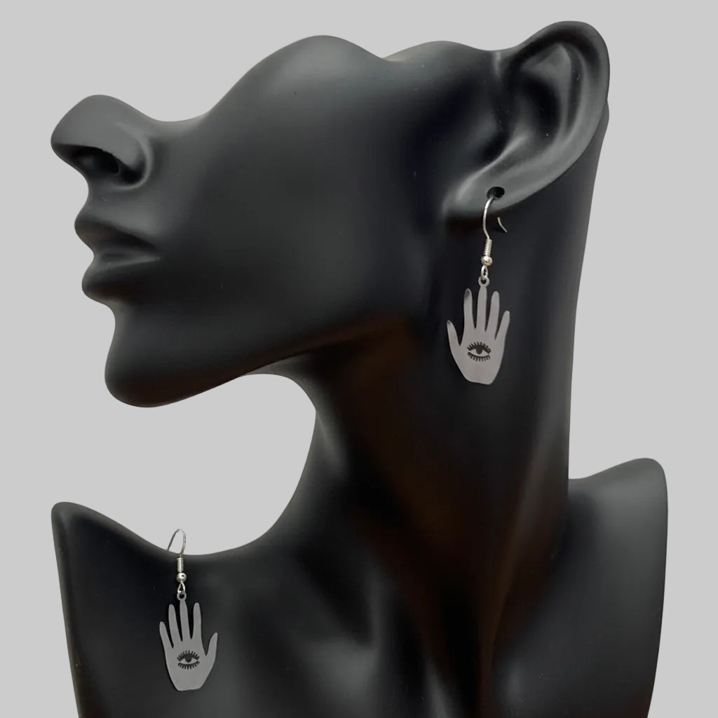 A pair of silver hamsa hand drop earrings with a small evil eye charm, displayed on a matte black female bust against a wrinkled white backdrop.