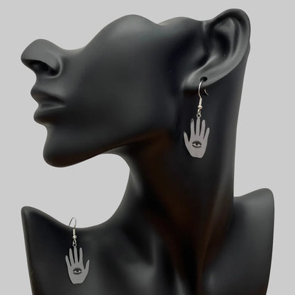 A pair of silver hamsa hand drop earrings with a small evil eye charm, displayed on a matte black female bust against a wrinkled white backdrop.
