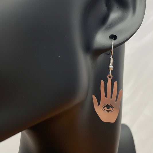 A single dangling earring displayed on a black mannequin head against a light fabric backdrop. Hand-shaped charm with a small black eye detail.