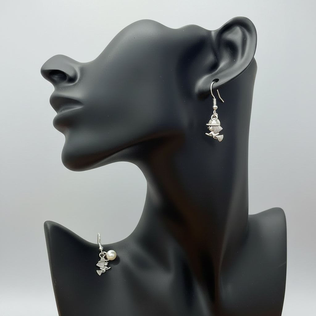 A pair of silver-toned dangle earrings with small witch charms and a pearl accent, displayed on a black mannequin bust against a light gray backdrop.