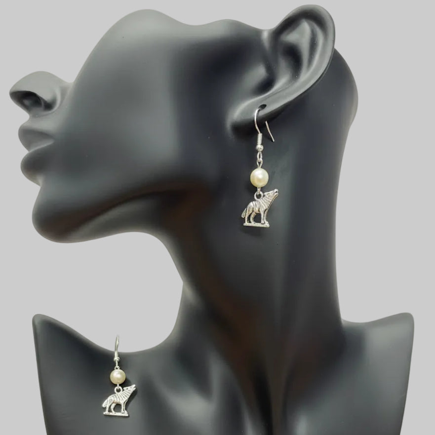 A pair of silver-toned dangle earrings with small animal charms and white pearl-like beads, displayed on a black mannequin bust against a light backdrop