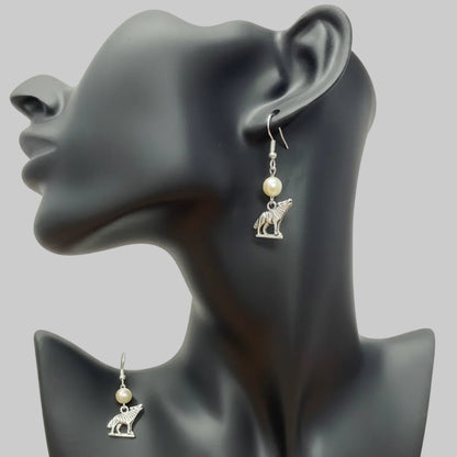 A pair of silver-toned dangle earrings with small animal charms and white pearl-like beads, displayed on a black mannequin bust against a light backdrop