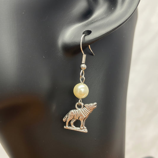 A silver-toned dangle earring displayed on a black mannequin ear. The earring features a small silver wolf charm and a round pale resin pearl.