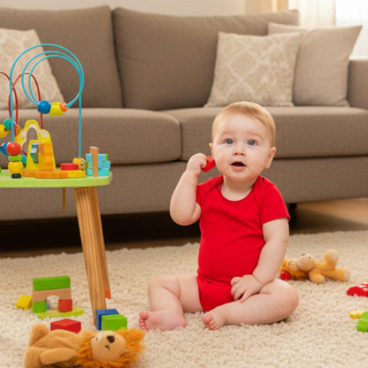 Baby in a red bodysuit sitting next to a colorful play table 
