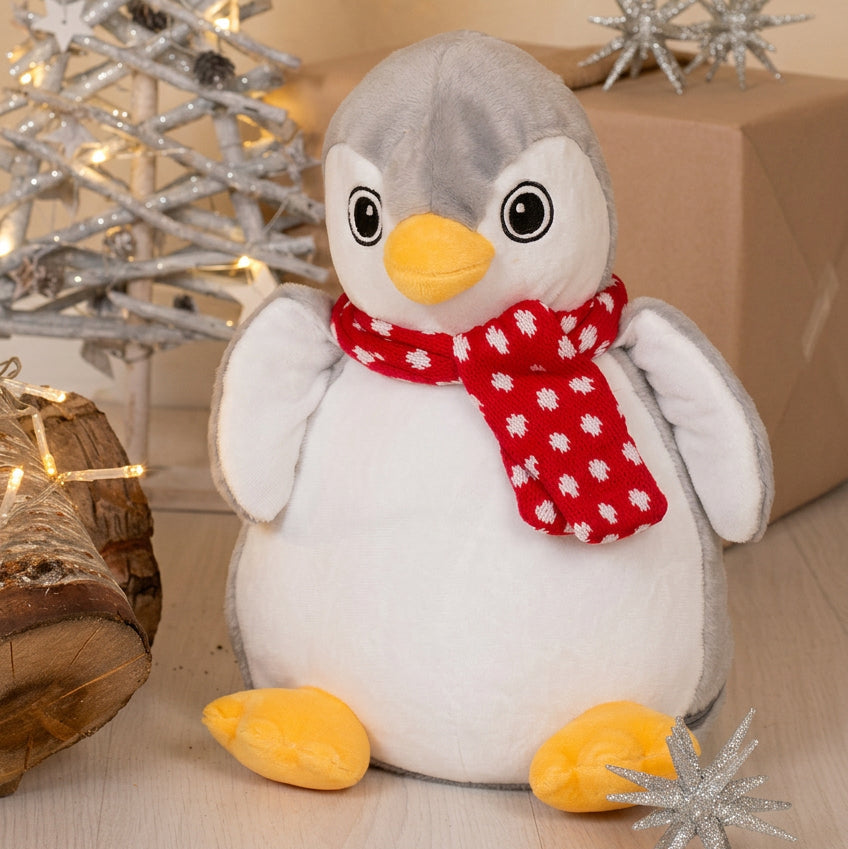 A plush penguin toy with a gray head, white body, yellow beak and feet, and a red scarf with white snowflake pattern, posed on a light wood surface with holiday decor in the background.