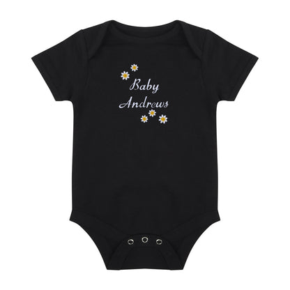 A black short-sleeve baby bodysuit with 'Baby Andrews' embroidered in white script on the chest, decorated with small white and yellow daisy flowers. The bodysuit has three silver metal snap buttons at the crotch and is displayed flat against a solid white background.