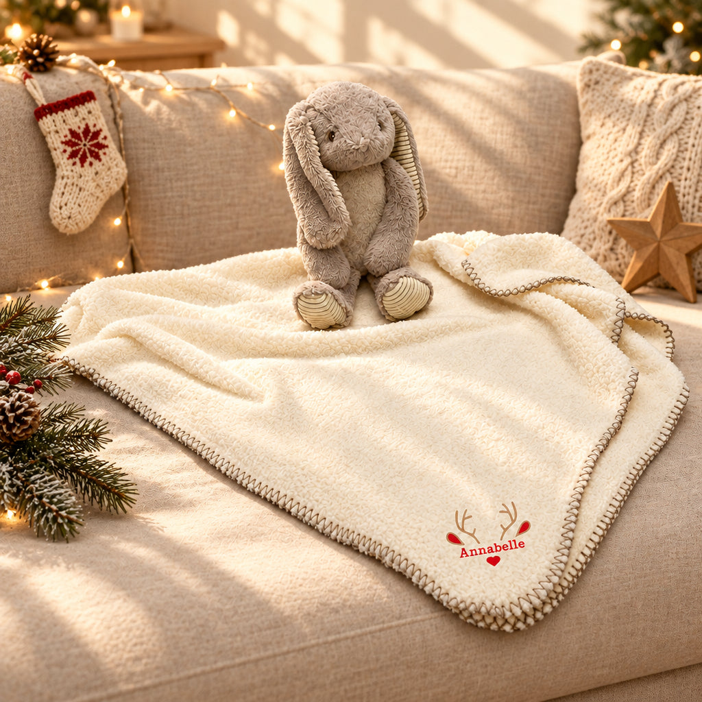 A cream sherpa blanket with a knitted border and personalized red embroidery reading 'Annabelle' with reindeer antlers, draped over a beige sofa with a plush bunny toy, holiday lights, pine branches, and a knitted stocking in the background.