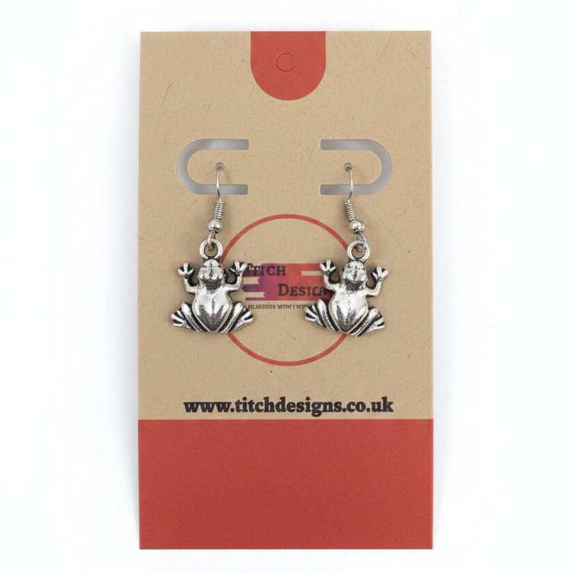 A pair of silver-toned frog dangle earrings displayed on a kraft paper card with red accents.