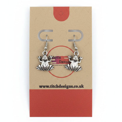 A pair of silver-toned frog dangle earrings displayed on a kraft paper card with red accents.