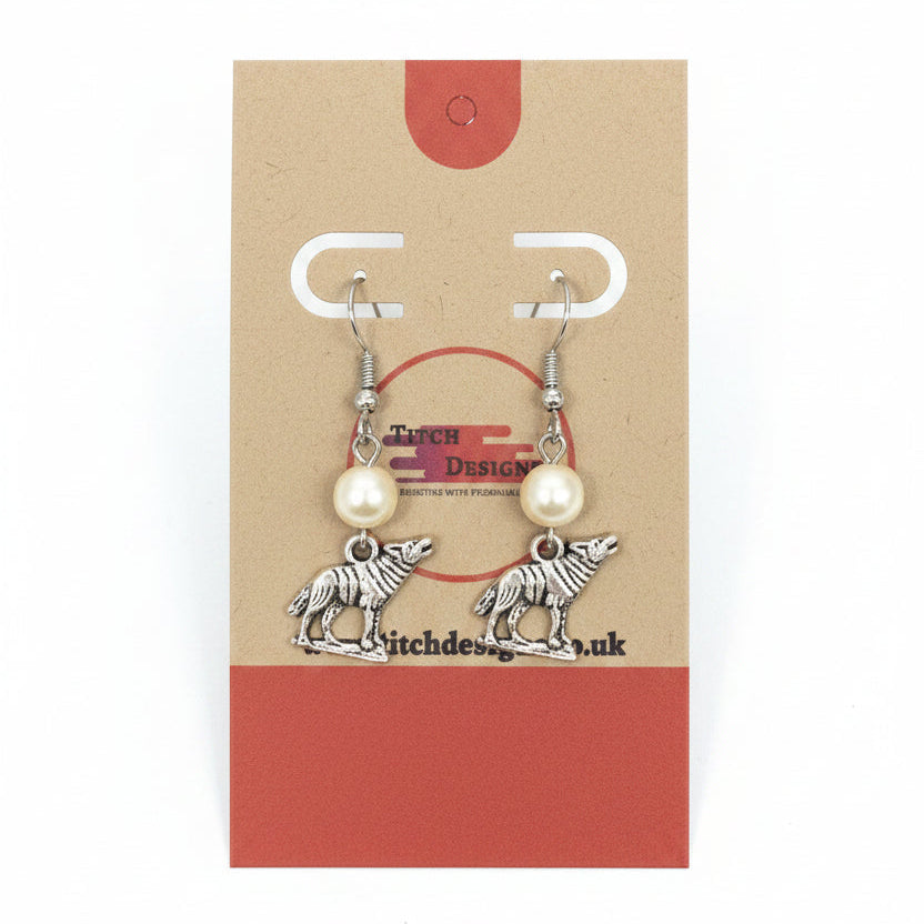 A pair of handmade dangle earrings displayed on a brown kraft paper card. The earrings feature silver-toned metal hooks and pendants shaped like wolves, with small white pearl-like beads above each wolf charm.