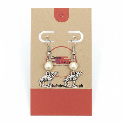 A pair of handmade dangle earrings displayed on a brown kraft paper card. The earrings feature silver-toned metal hooks and pendants shaped like wolves, with small white pearl-like beads above each wolf charm.