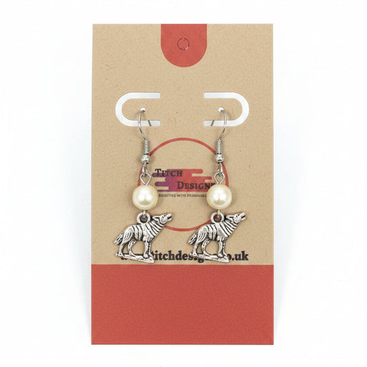 A pair of handmade dangle earrings displayed on a brown kraft paper card. The earrings feature silver-toned metal hooks and pendants shaped like wolves, with small white pearl-like beads above each wolf charm.