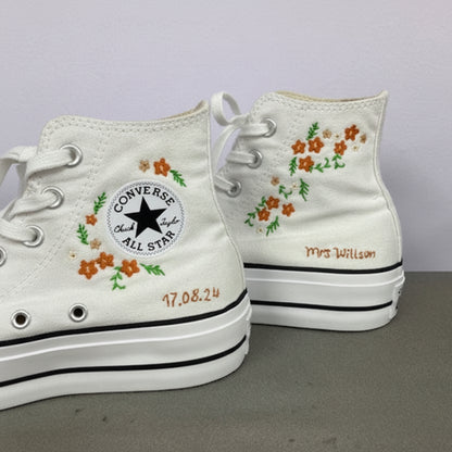 A pair of white Converse high-top sneakers with custom embroidery. The left shoe features an embroidered floral pattern around the ankle patch, and the right shoe has matching floral embroidery on the side, along with personalized text