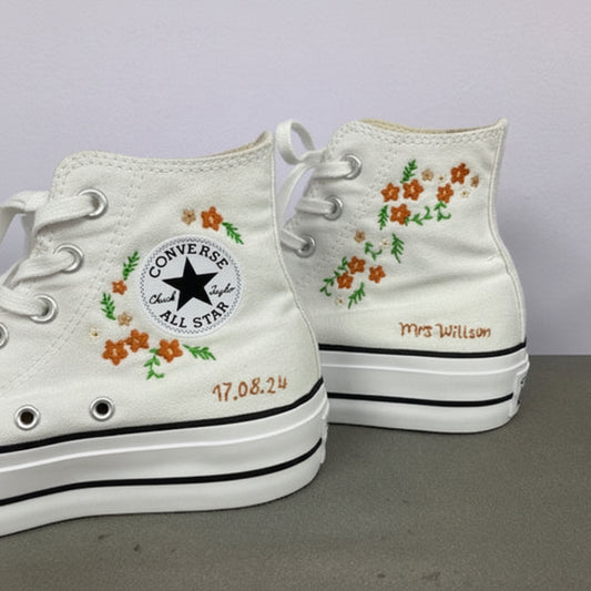A pair of white Converse high-top sneakers with custom embroidery. The left shoe features an embroidered floral pattern around the ankle patch, and the right shoe has matching floral embroidery on the side, along with personalized text