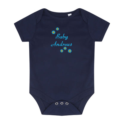 A navy blue short-sleeve baby bodysuit with 'Baby Andrews' embroidered in light blue script on the chest, decorated with small blue and yellow flowers. The bodysuit features three silver snap closures at the crotch and is displayed flat against a solid white background.