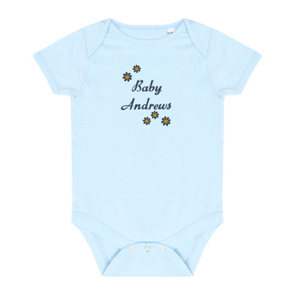 A light blue short-sleeve baby bodysuit with 'Baby Andrews' embroidered in navy script on the chest, surrounded by small yellow and navy flower motifs. The bodysuit has three white snap buttons at the crotch and is displayed flat against a solid white background.
