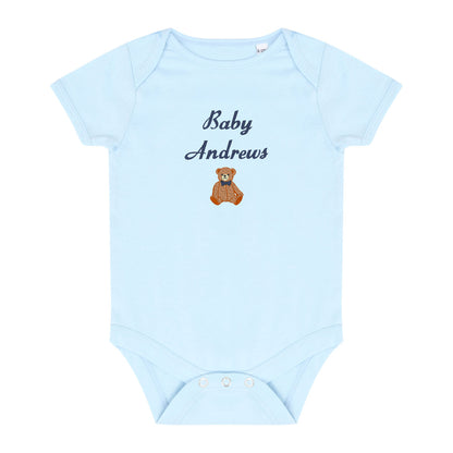 A light blue short-sleeve baby bodysuit with 'Baby Andrews' embroidered in navy script and a small brown teddy bear graphic on the chest, shown on a plain white background.