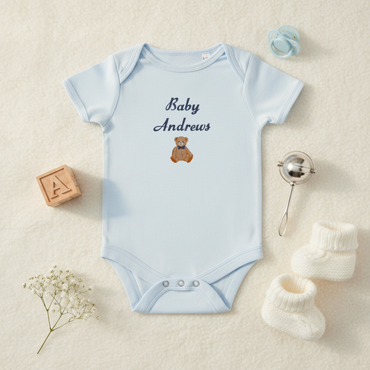 A light blue baby bodysuit with 'Baby Andrews' and a teddy bear graphic embroidered on the chest, styled in a flat lay with baby accessories on a cream textured background.