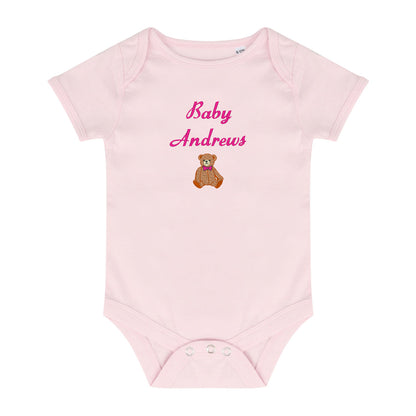 A light pink short-sleeve baby bodysuit with 'Baby Andrews' embroidered in pink script on the chest, above a small brown teddy bear wearing a pink bow tie. The bodysuit has three snap closures at the crotch and is shown flat against a white background.