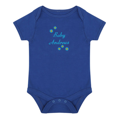 A royal blue short-sleeve baby bodysuit with 'Baby Andrews' embroidered in light blue script on the chest, decorated with small yellow and blue flower graphics, shown on a plain white background.