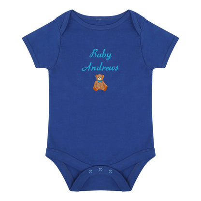 A royal blue short-sleeve baby bodysuit with 'Baby Andrews' embroidered in light blue script above a small brown teddy bear wearing a blue bow tie, shown on a plain white background.