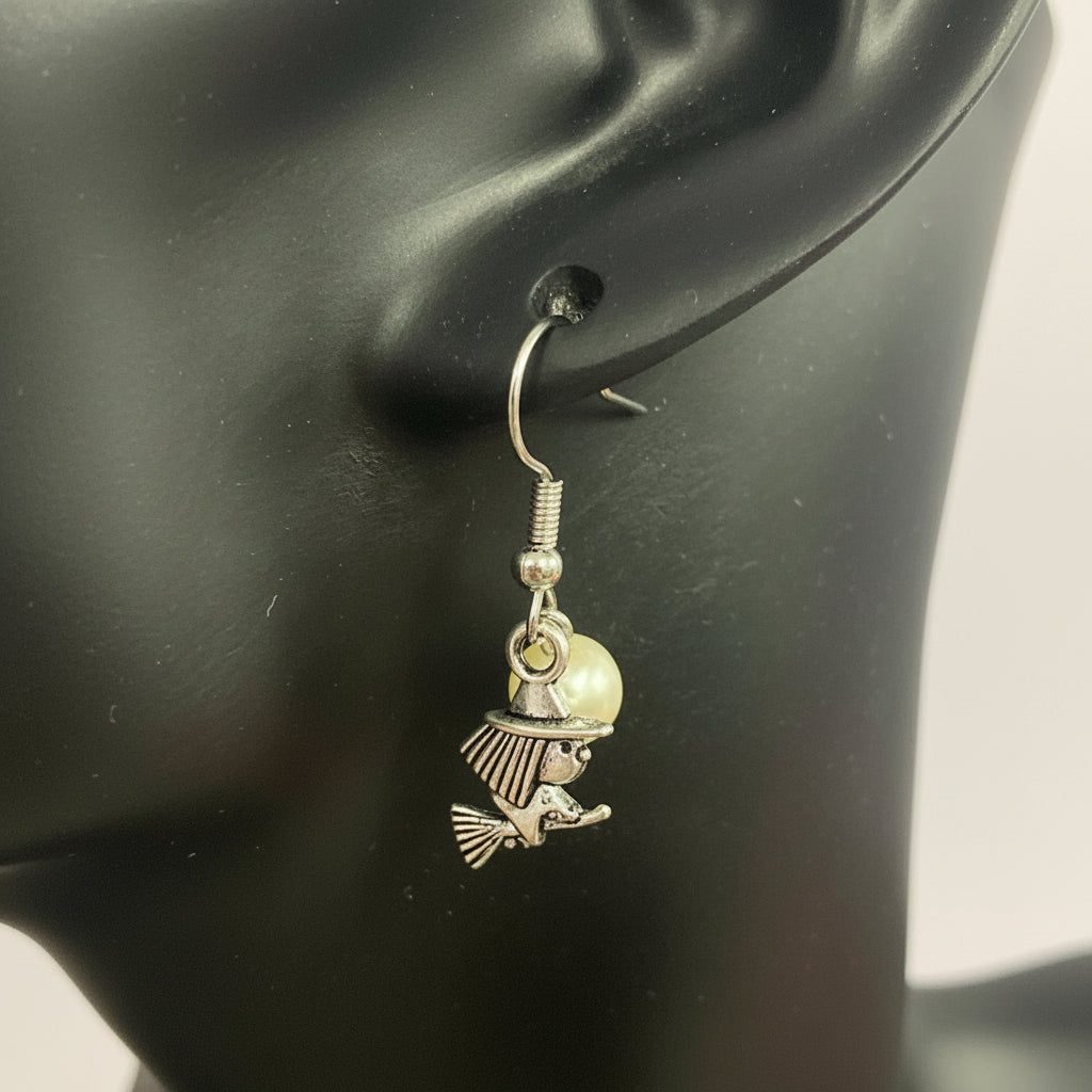 A silver witch charm dangle earring with a small pearl bead, shown on a black mannequin ear against a light background.
