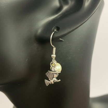 A silver witch charm dangle earring with a small pearl bead, shown on a black mannequin ear against a light background.