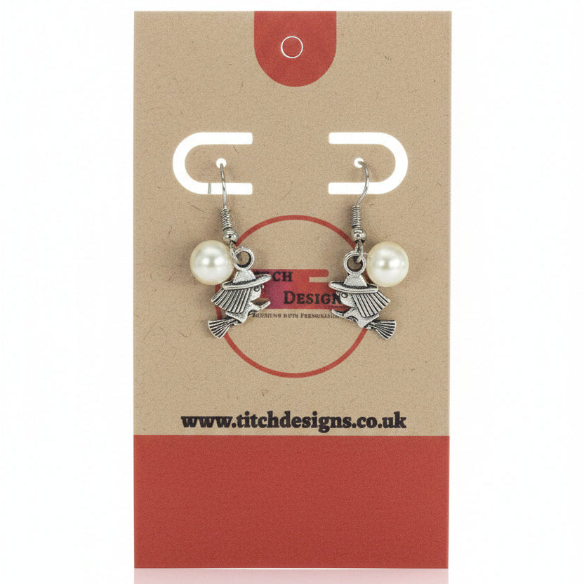 A pair of silver-toned witch charm earrings with white pearl beads, displayed on a brown kraft paper card with red accents. The card features the brand name 'Titch Designs'