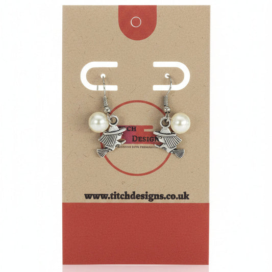 A pair of silver-toned witch charm earrings with white pearl beads, displayed on a brown kraft paper card with red accents. The card features the brand name 'Titch Designs'