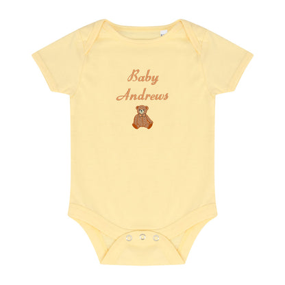 A pale yellow short-sleeve baby bodysuit with 'Baby Andrews' embroidered in a brown cursive font above a small brown teddy bear graphic, shown on a plain white background.