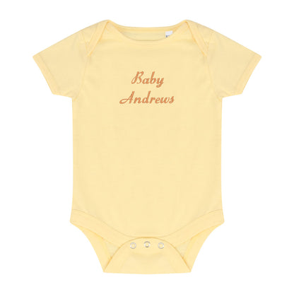 A pale yellow short-sleeve baby bodysuit with 'Baby Andrews' embroidered in a rose-gold cursive font on the chest, displayed flat against a solid white background.